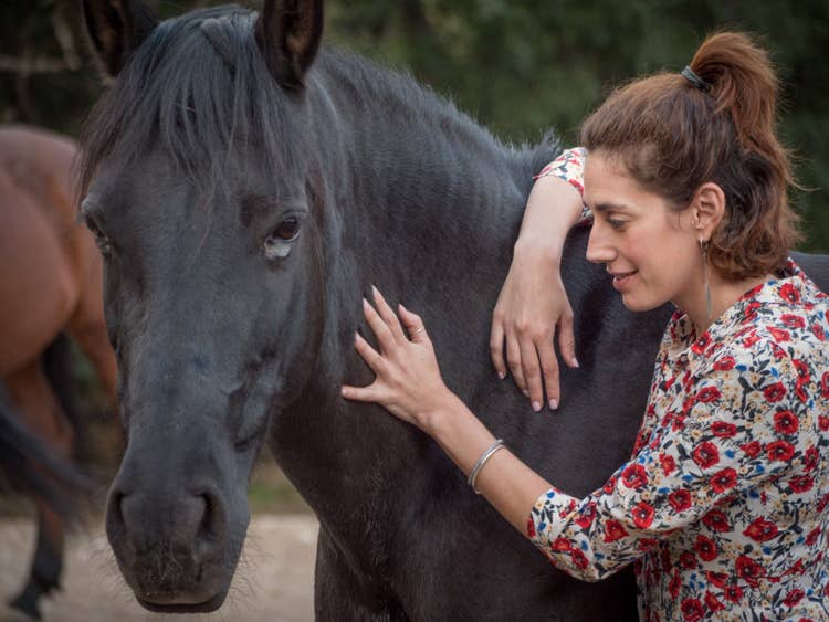 A female rider in a floral shirt soothingly stoking a black horse's neck.